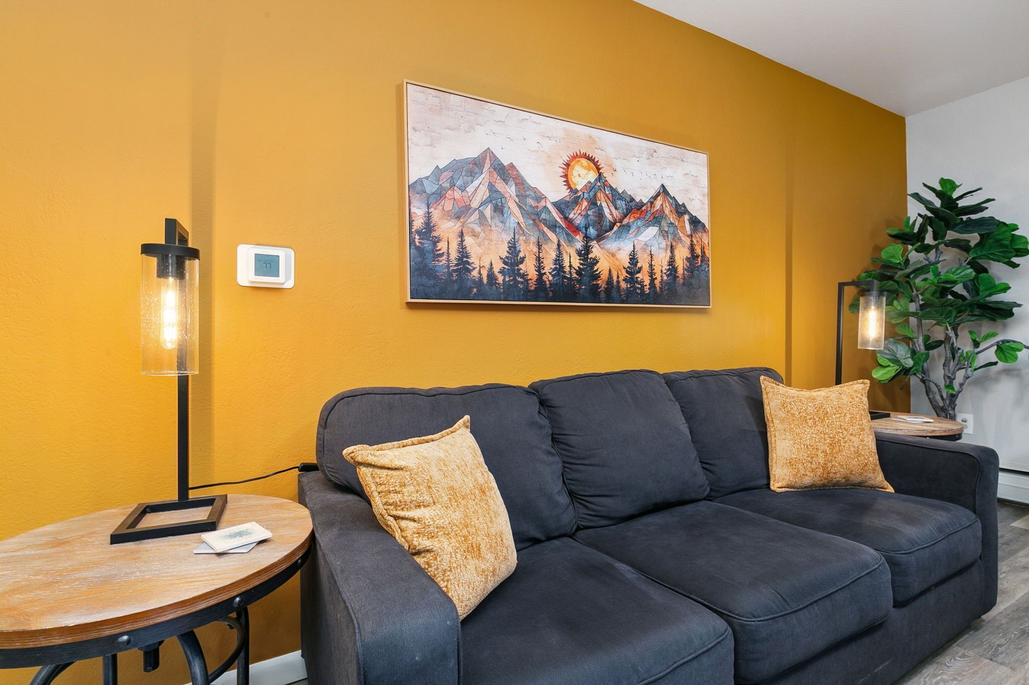 A modern living room with a black sofa, two cushions, a round wooden table, a floor lamp, and a painting of mountains on the wall.