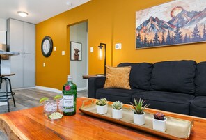Una sala de estar con un sofá negro, una mesa de centro de madera con una bandeja que contiene bebidas y plantas, y un cuadro de montañas en la pared.