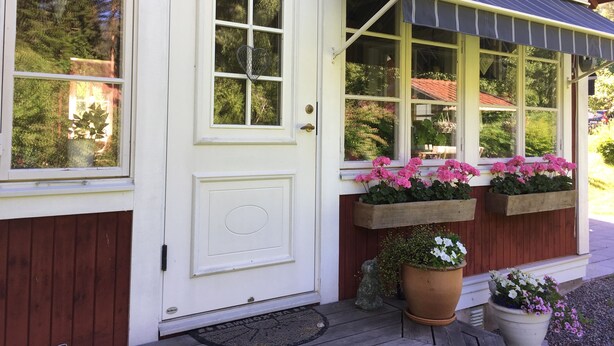 Une porte blanche avec une fenêtre, un porche doté d’un auvent à rayures et des plantes en pot ornées de fleurs roses.