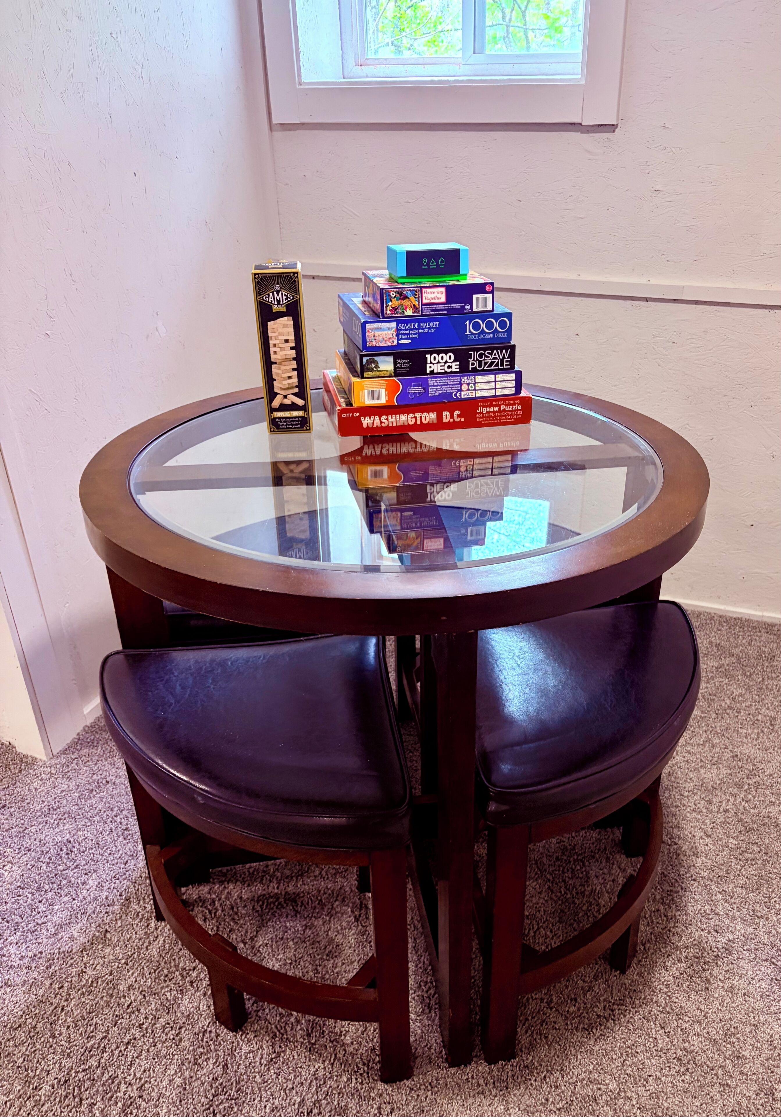 Ein runder Glastisch mit dunklem Holzfuß, umgeben von zwei dunklen Holzstühlen. Auf dem Tisch befinden sich verschiedene Puzzleboxen, darunter ein Puzzle mit 1000 Teilen und ein Puzzle zum Thema 'Washington D.C.'.