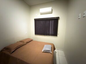 A single bed with a brown bedspread and pillows, an air conditioning unit above the window, and a tiled floor. - CONDOMÍNIO SONHARE - 204 (Anapolis)
