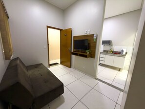 A compact living space with a sofa, a television mounted on a wooden panel, and a kitchenette area. - CONDOMÍNIO SONHARE - 204 (Anapolis)