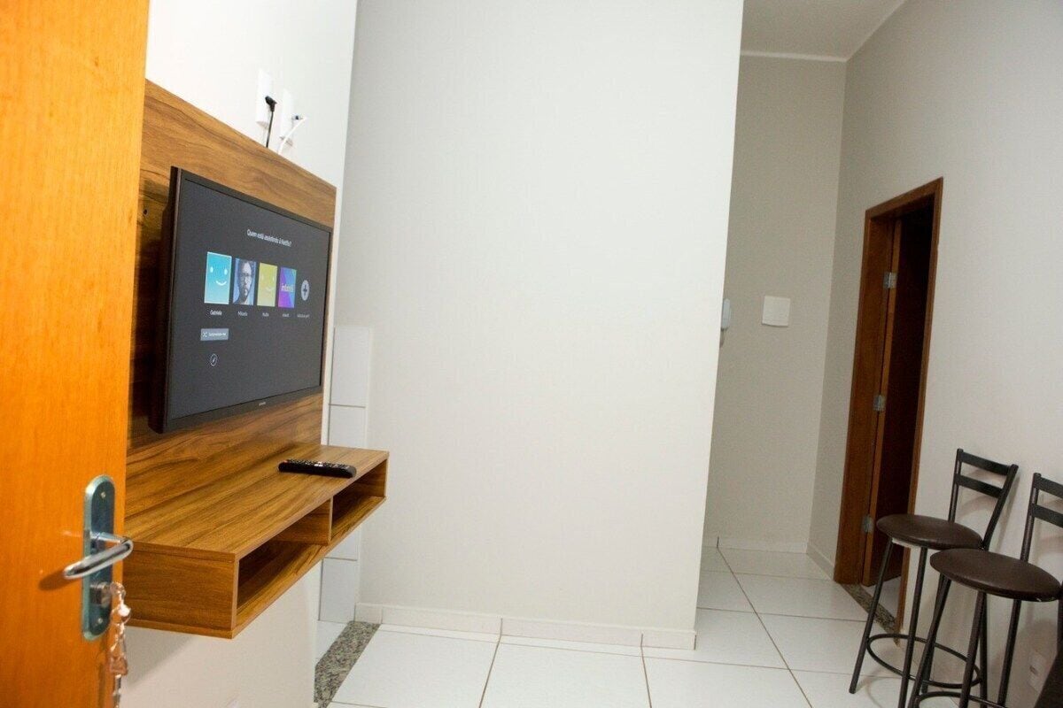 Una habitación con un televisor de pantalla plana montado en un estante de madera, un control remoto sobre el estante y dos taburetes de bar pegados a la pared.