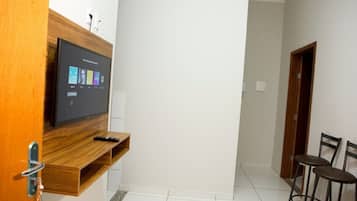 Una habitación con un televisor de pantalla plana montado en un estante de madera, un control remoto sobre el estante y dos taburetes de bar pegados a la pared.