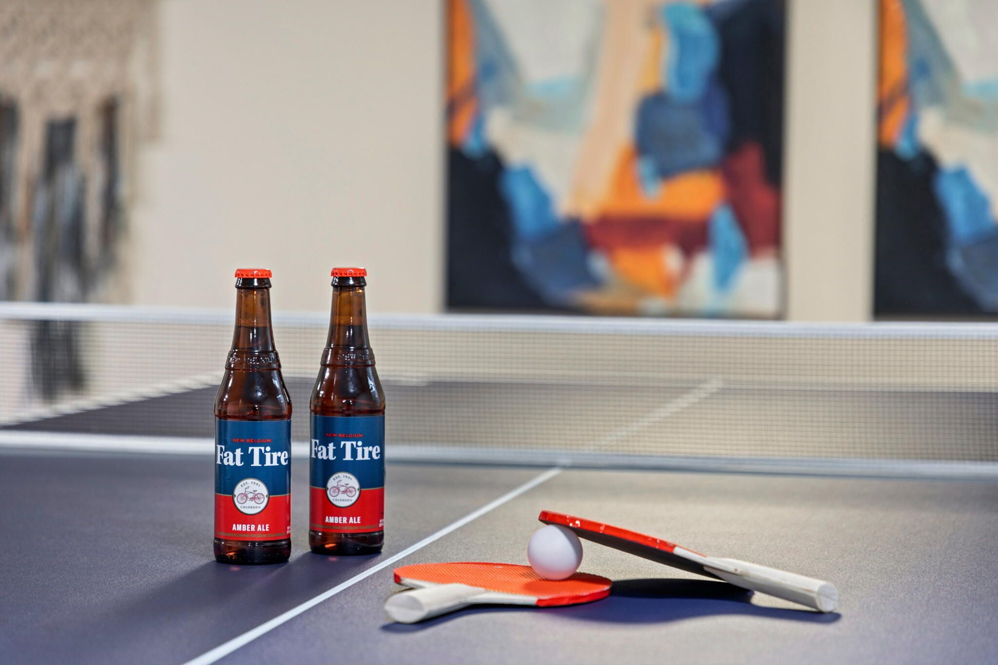 Two bottles of Fat Tire Amber Ale on a ping pong table with a paddle and ball.
