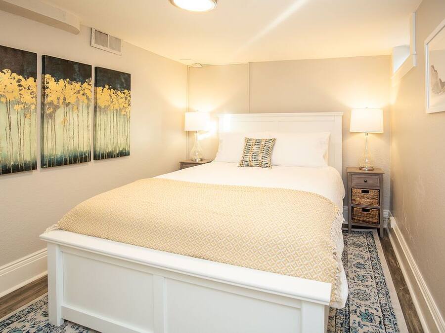 Un dormitorio con una cama blanca, dos lámparas de noche, un tapete estampado y tres cuadros colgados en la pared.