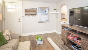 A living room with a sofa, a coffee table with a plant, a TV stand with books, a painting on the wall, and a door with a window. - Cozy Retreat | Walk to Unc | Free Cruiser Bikes (Greeley)