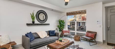 A modern living room with a grey sofa, a wooden coffee table, and a cowhide rug. There is a ceiling fan, a window with blinds, and a door.