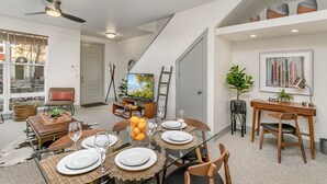 Un'area pranzo con tavolo in vetro apparecchiato per quattro persone, circondato da sedie in legno. Si vede una ventola a soffitto e una scala che conduce a un altro livello.