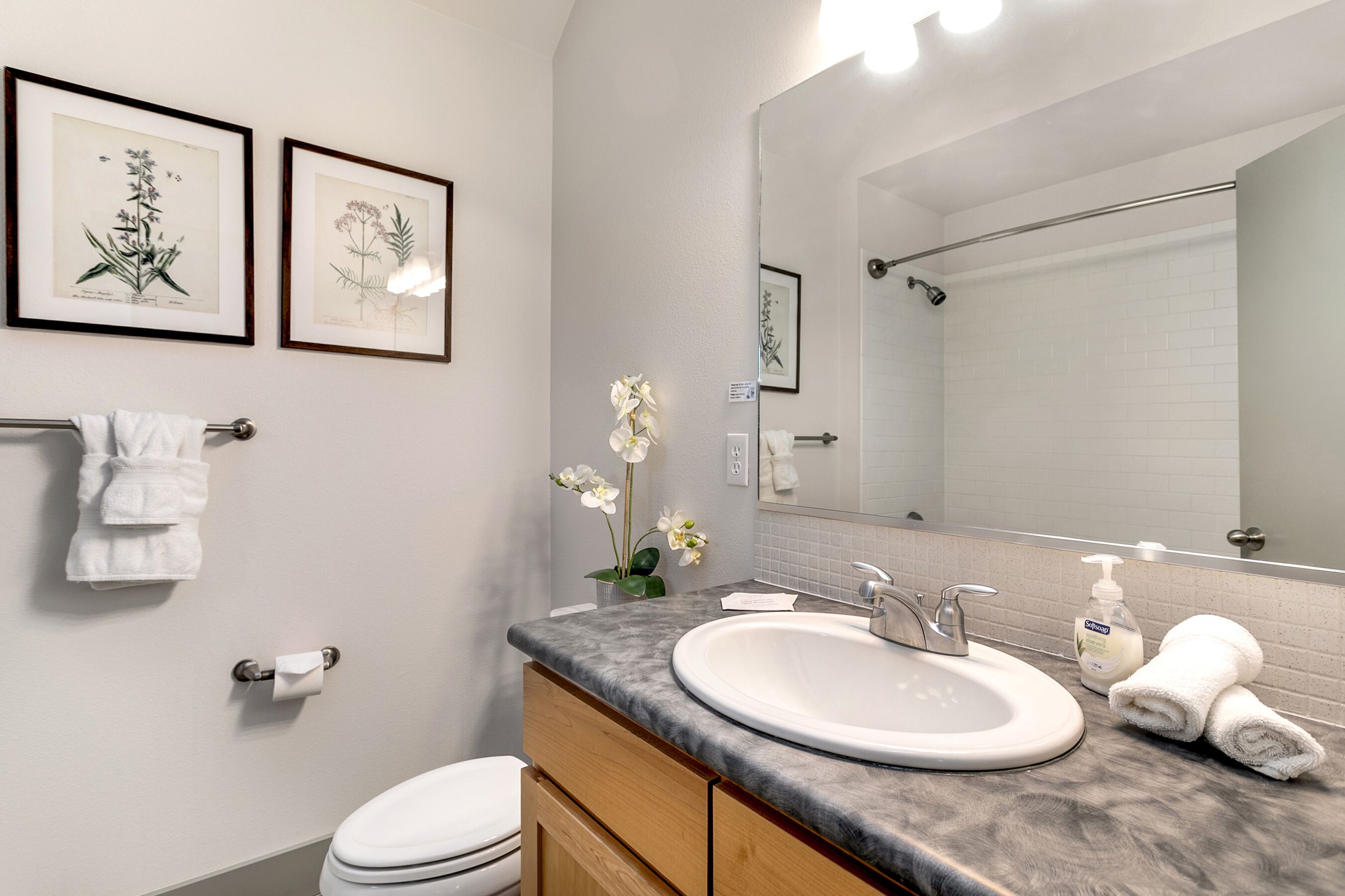 A modern bathroom with a white sink, vanity, and toilet. There are framed botanical prints on the wall, a towel rack, and a mirror above the sink.