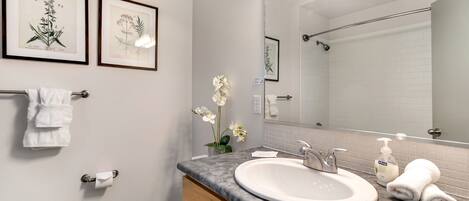 A modern bathroom with a white sink, vanity, and toilet. There are framed botanical prints on the wall, a towel rack, and a mirror above the sink.