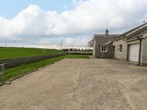 Une allée de gravier mène à un bâtiment en pierre doté d’un garage, qui est entouré d’un champ herbeux et d’un petit muret en pierre.