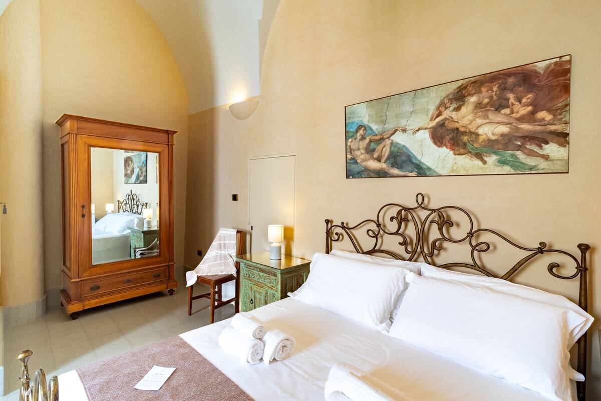 A hotel room with a bed, a wooden wardrobe, a nightstand with a lamp, and a painting on the wall.