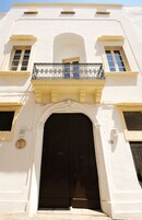 Un edificio blanco con una puerta marrón oscuro, un balcón con barandilla metálica y una ventana pequeña con marco azul.