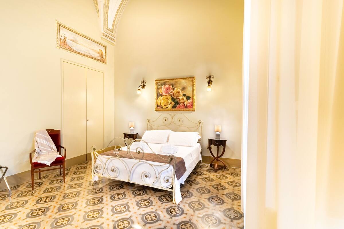 A bedroom with a bed, a chair, a painting on the wall, and patterned flooring.