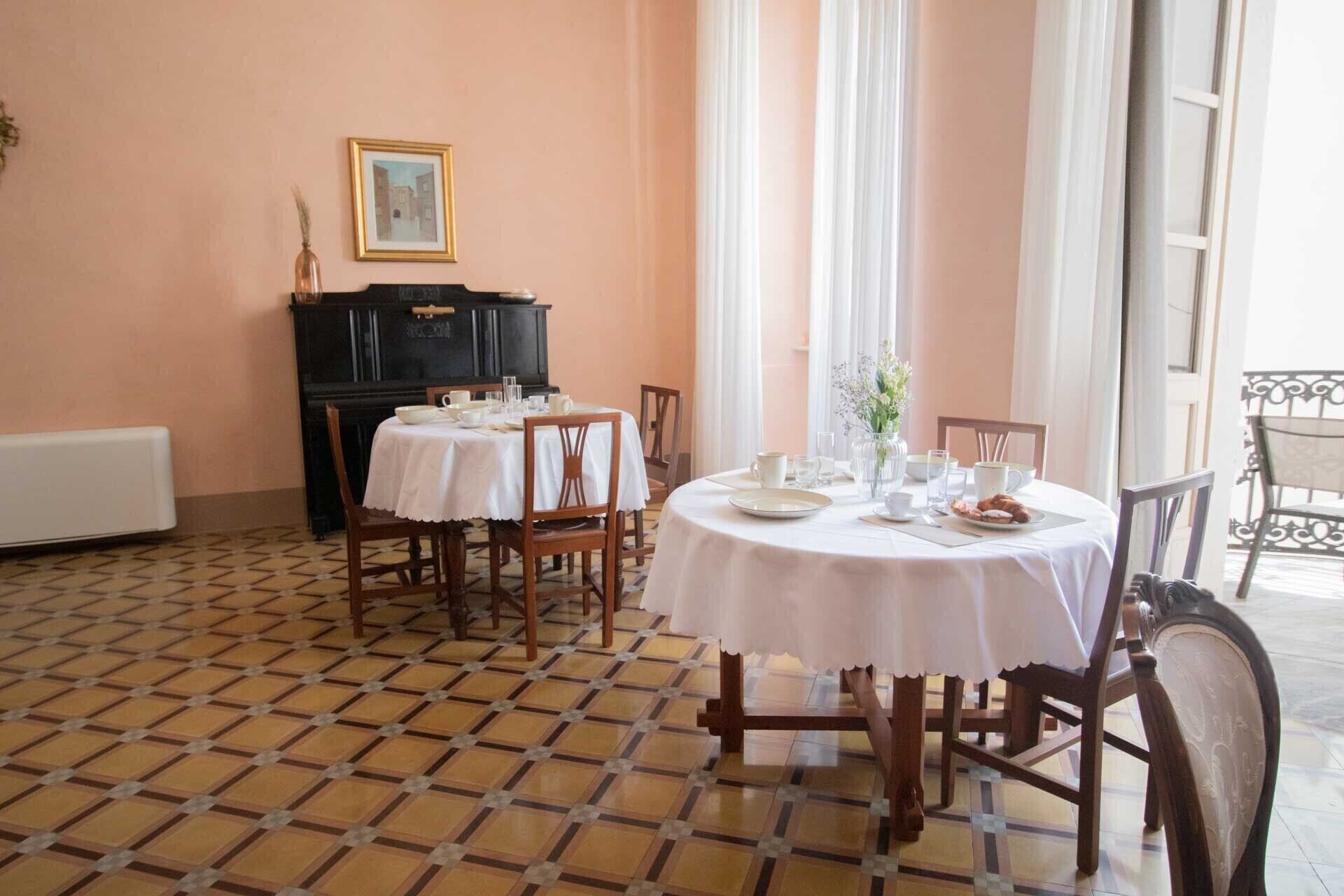 Un espace repas avec des tables rondes disposées pour accueillir quatre personnes, chacune étant recouverte d’une nappe blanche et entourée de chaises en bois. On y trouve un buffet noir sur lequel est posée une image encadrée, ainsi qu’un radiateur fixé au mur.