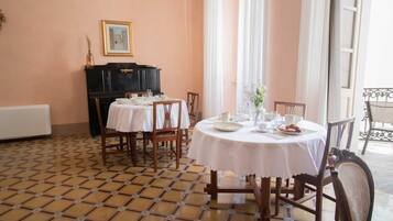 Un espace repas avec des tables rondes disposées pour accueillir quatre personnes, chacune étant recouverte d’une nappe blanche et entourée de chaises en bois. On y trouve un buffet noir sur lequel est posée une image encadrée, ainsi qu’un radiateur fixé au mur.