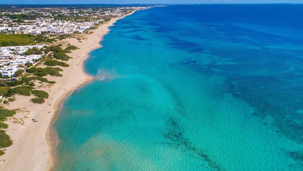 An aerial view of a coastal area with a sandy beach, clear turquoise waters, and a residential area with white buildings. - 3150 Villa Giovì - Appartamento 2 by Barbarhouse (Manduria)