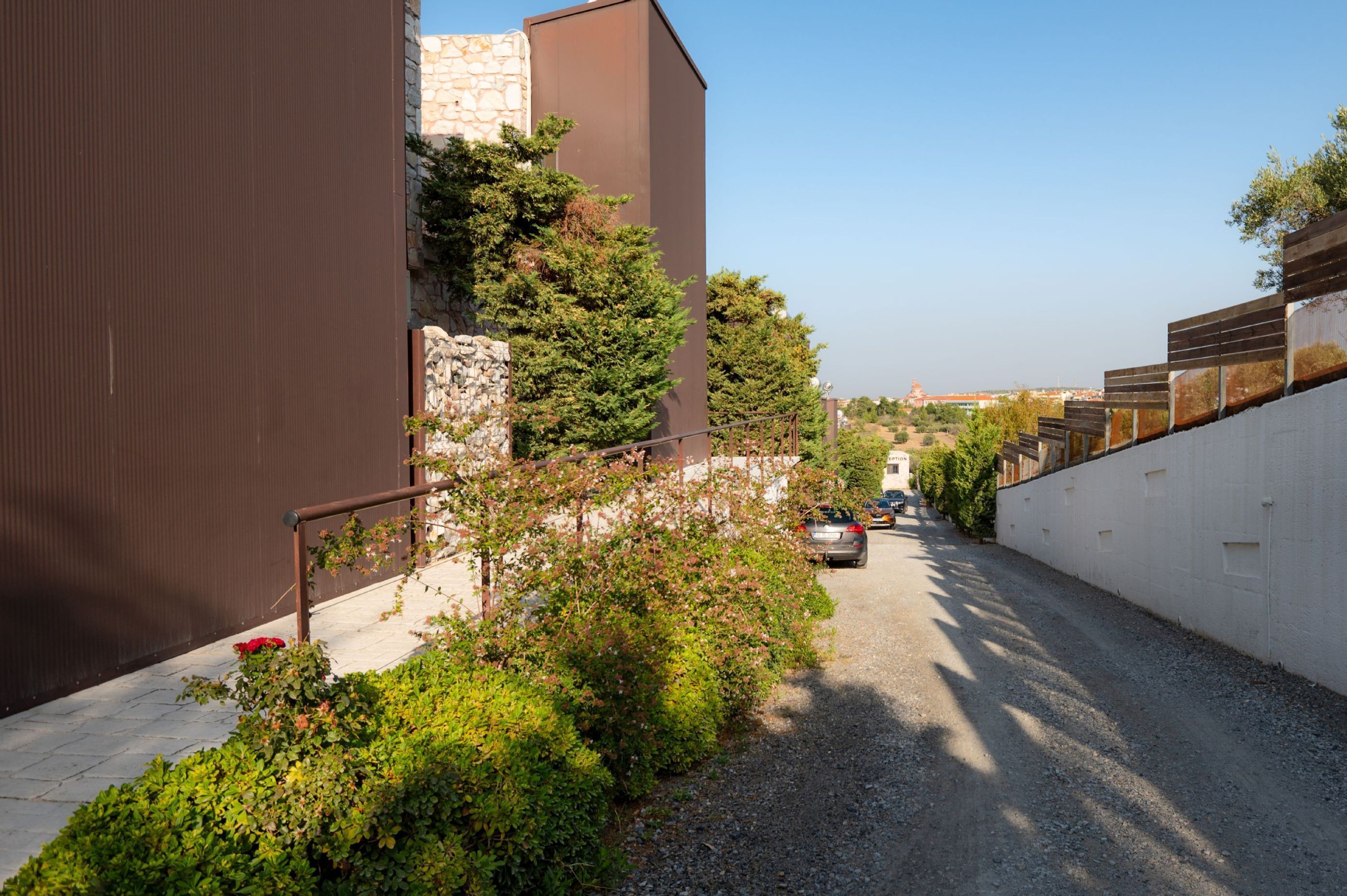 Una calle estrecha con un camino de grava, un coche estacionado y un edificio de fachada de piedra junto a una estructura moderna en tonos marrones.