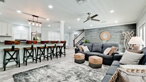 Eine moderne Küche mit weißen Schränken, einer Bar mit Hockern, einem grauen Sofa mit Kissen und einer Wanddekoration.