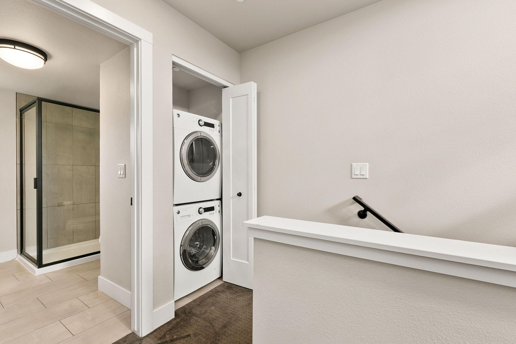 A laundry room with two stacked front-loading washing machines, a glass shower enclosure, and a white bench.