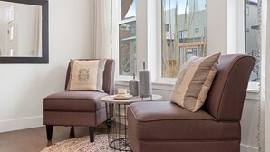Ett modernt vardagsrum med två bruna soffor, ett litet runt bord och utsikt över byggnader genom stora fönster.