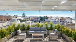 A rooftop terrace with a white sofa, chairs, and planters, overlooking a coastal cityscape with buildings and the sea. - 3135 La Voce del Mare by Barbarhouse (Nardò)