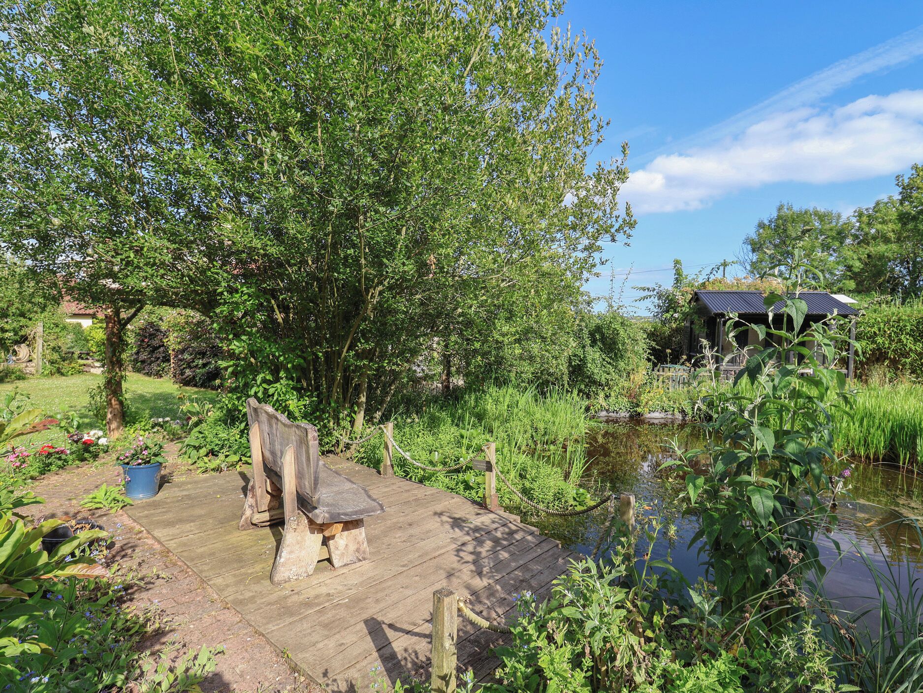 A wooden deck with a rustic bench, surrounded by lush greenery and a pond.