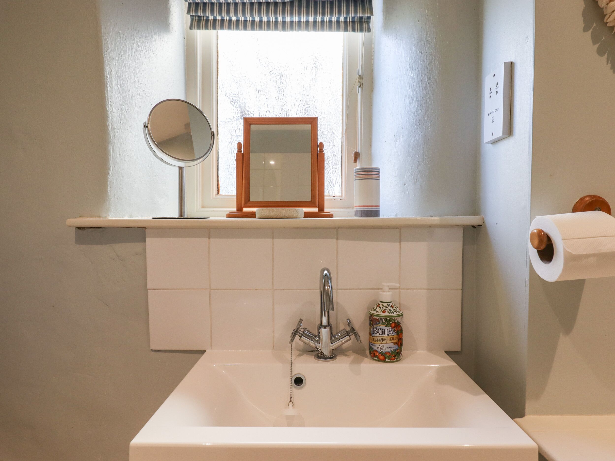 Ein Badezimmer mit weißem Waschbecken, einem Spiegel, einem Fenster mit gestreifter Jalousie und einem Toilettenpapierhalter.