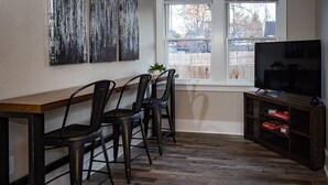 Une cuisine moderne avec une table en bois, deux chaises noires, un téléviseur posé sur un meuble en bois et une fenêtre donnant sur une clôture.