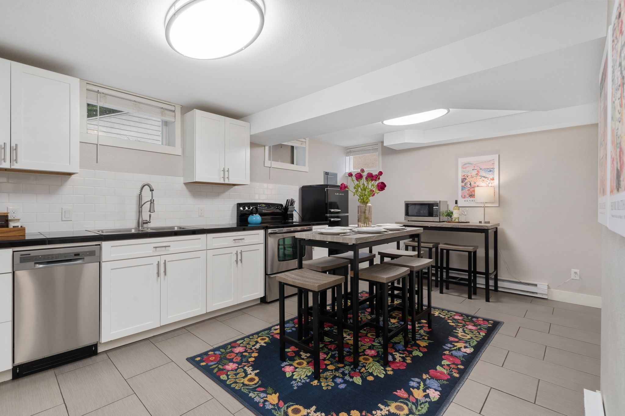 A modern kitchen with white cabinets, a dishwasher, a refrigerator, a microwave, and a dining area with a floral rug.
