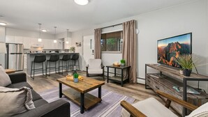 A modern apartment with a kitchen, dining area, and living room. It features a wooden coffee table, a sofa, a TV, and a large window with curtains. - Lavender Suite Relaxed, Walkable Old Town Stay (Fort Collins)
