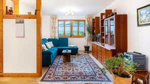 A living room with a blue sofa, a patterned rug, wooden cabinets, and a large window with a view of greenery. - Vacation apartment at Fuchsbühl  (Schopfheim)