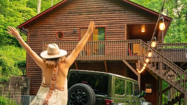 Une cabane en bois avec un escalier menant à un balcon, un VUS garé et une personne portant un chapeau et un short.
