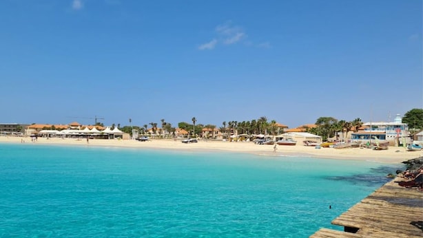 Una spiaggia costiera con acque turchesi cristalline, un molo di legno e edifici sullo sfondo con ombrelloni.