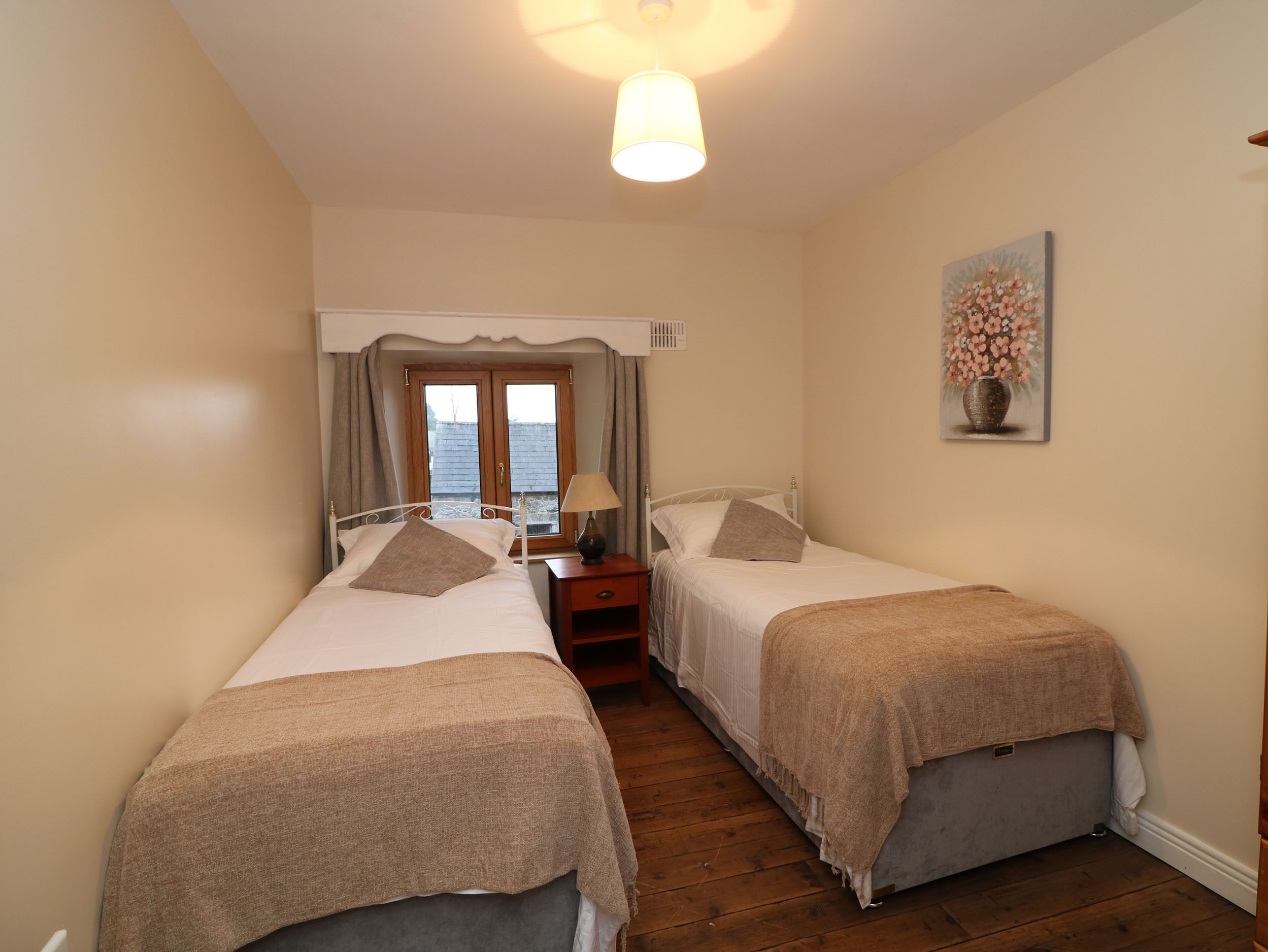 A bedroom with two single beds, a nightstand with a lamp, a window with curtains, and a painting on the wall.