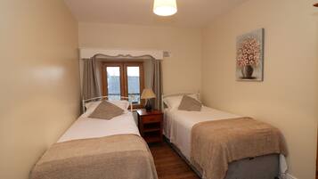 A bedroom with two single beds, a nightstand with a lamp, a window with curtains, and a painting on the wall.