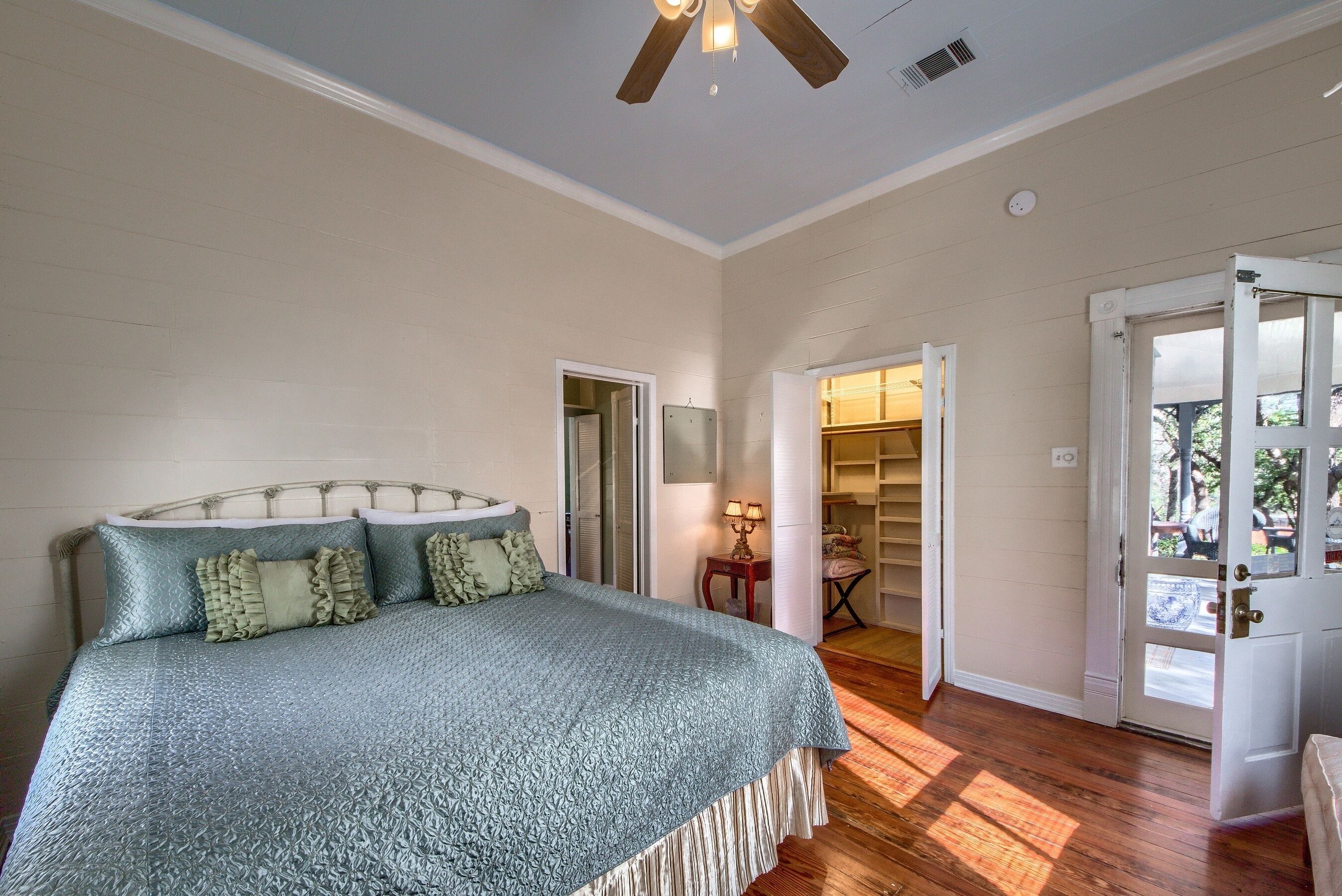 A bedroom with a bed, a ceiling fan, a door leading to a balcony, and a small table with a lamp.