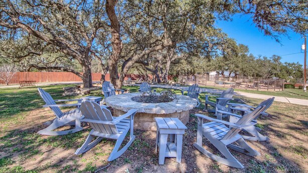 A circular stone fire pit surrounded by Adirondack chairs in a park setting.