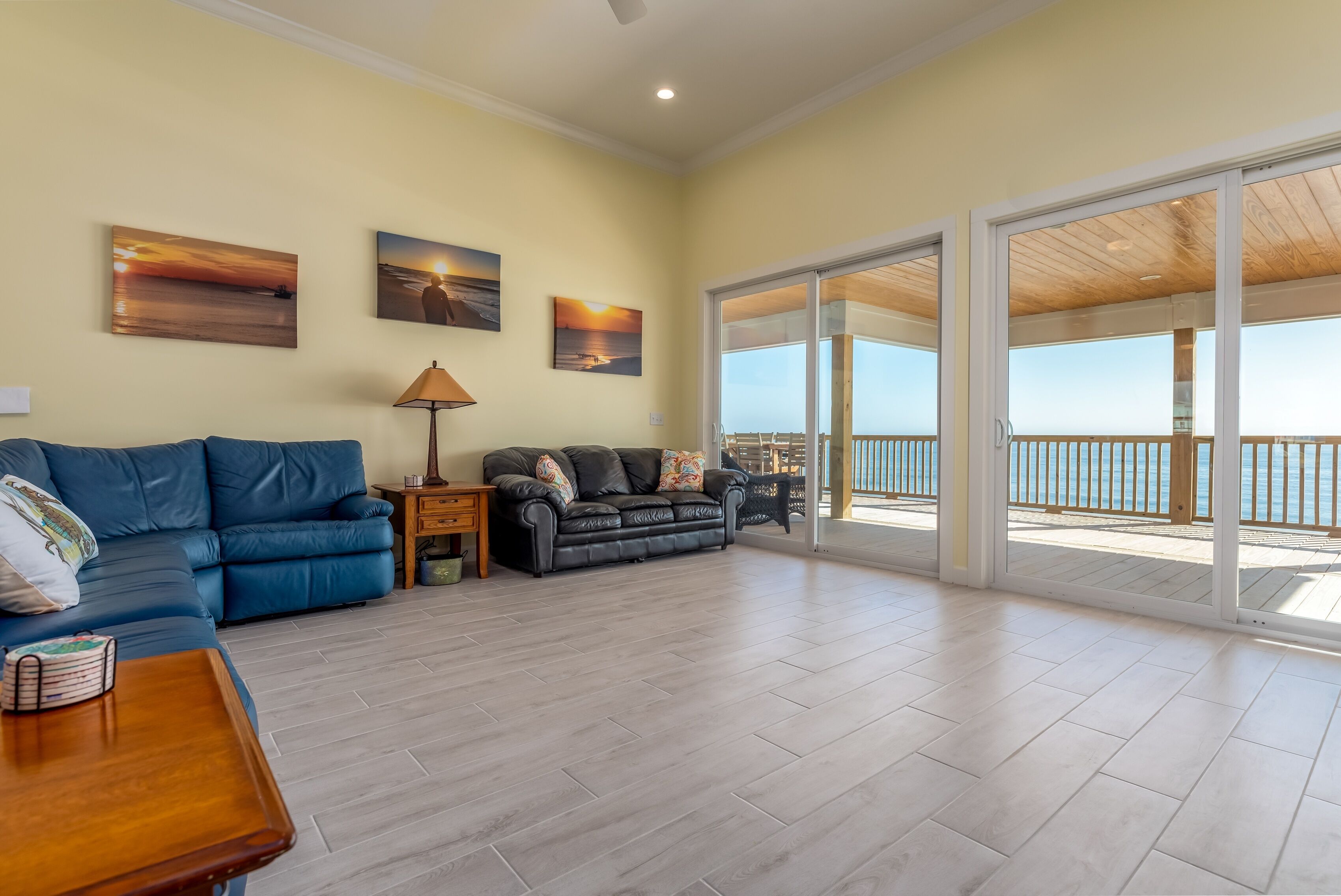 Uma sala de estar espaçosa com um sofá azul, uma mesa de centro de madeira e uma luminária. Há três quadros na parede e uma grande porta de vidro deslizante que dá para uma varanda com vista para o mar.