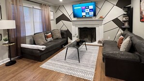 A modern living room with a fireplace, two sofas, a coffee table, and a flat-screen TV mounted above the fireplace. - 3 bedroom brick ranch (Apex)