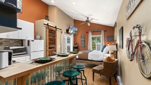 A compact kitchen with modern appliances, a bar area with stools, a bed with a view, and a bicycle mounted on the wall. - Industrial Old Town Bungalow W/ Free Cruiser Bikes (Fort Collins)