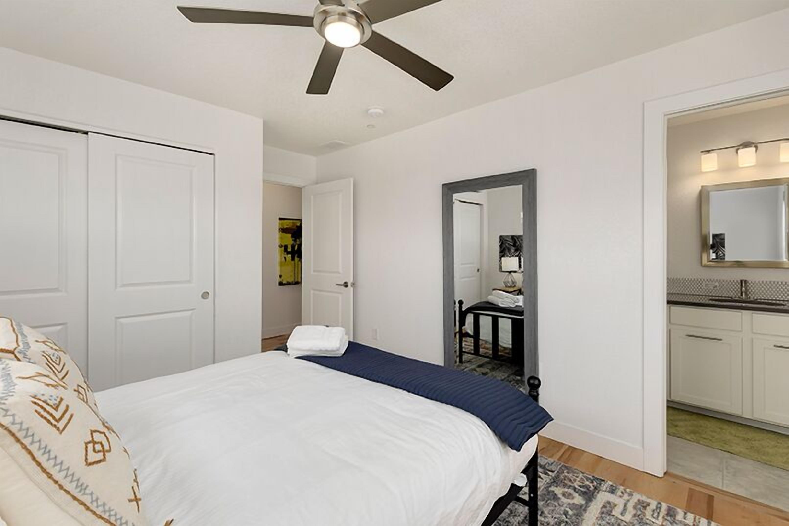A bedroom with a bed, a ceiling fan, a bathroom with a mirror, and a kitchen area.