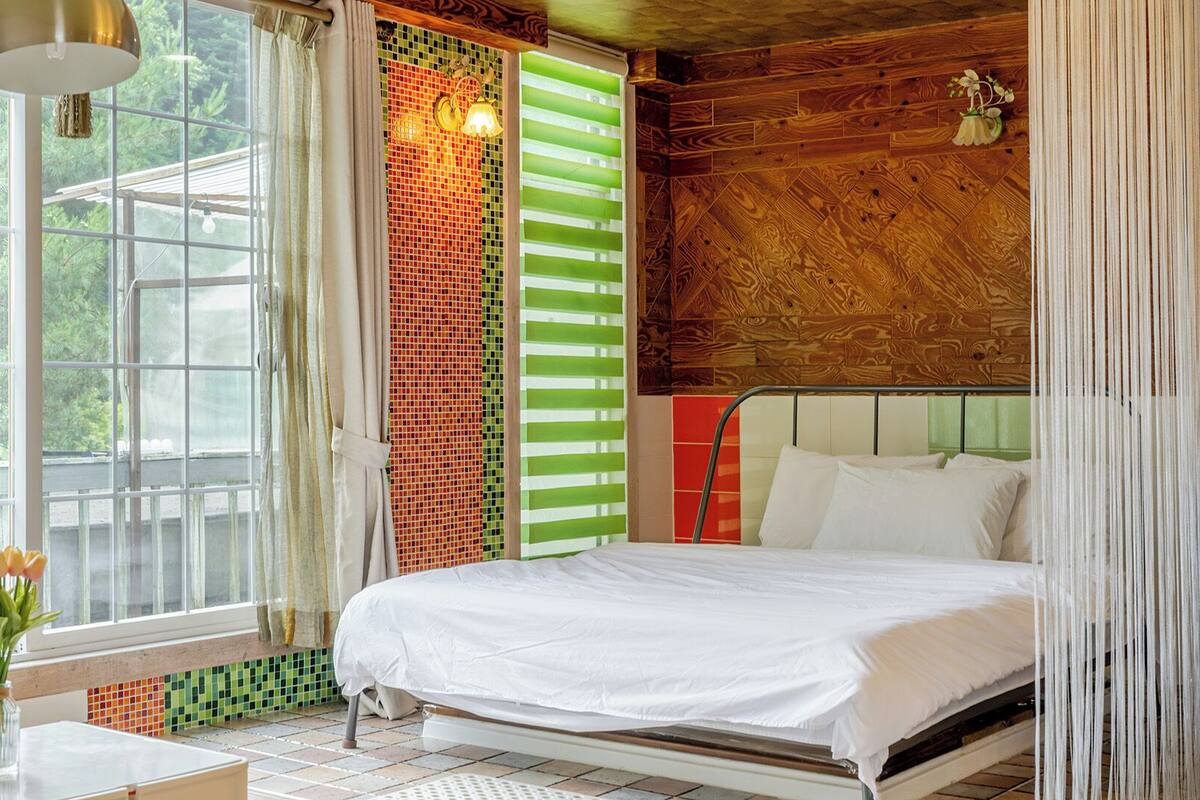 A bedroom with a bed, a window, a lamp, and a view of greenery outside.