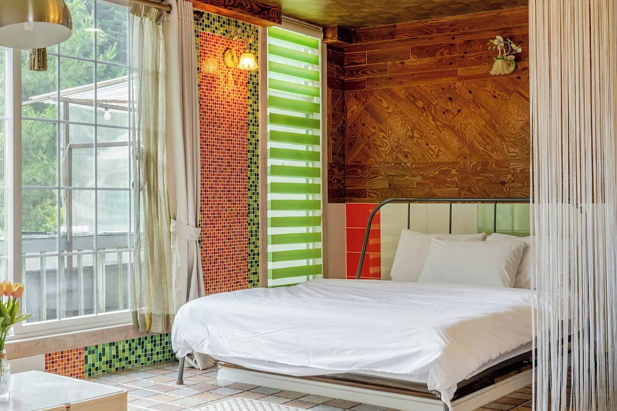Un dormitorio con cama, ventana, lámpara y vistas al verdor exterior.