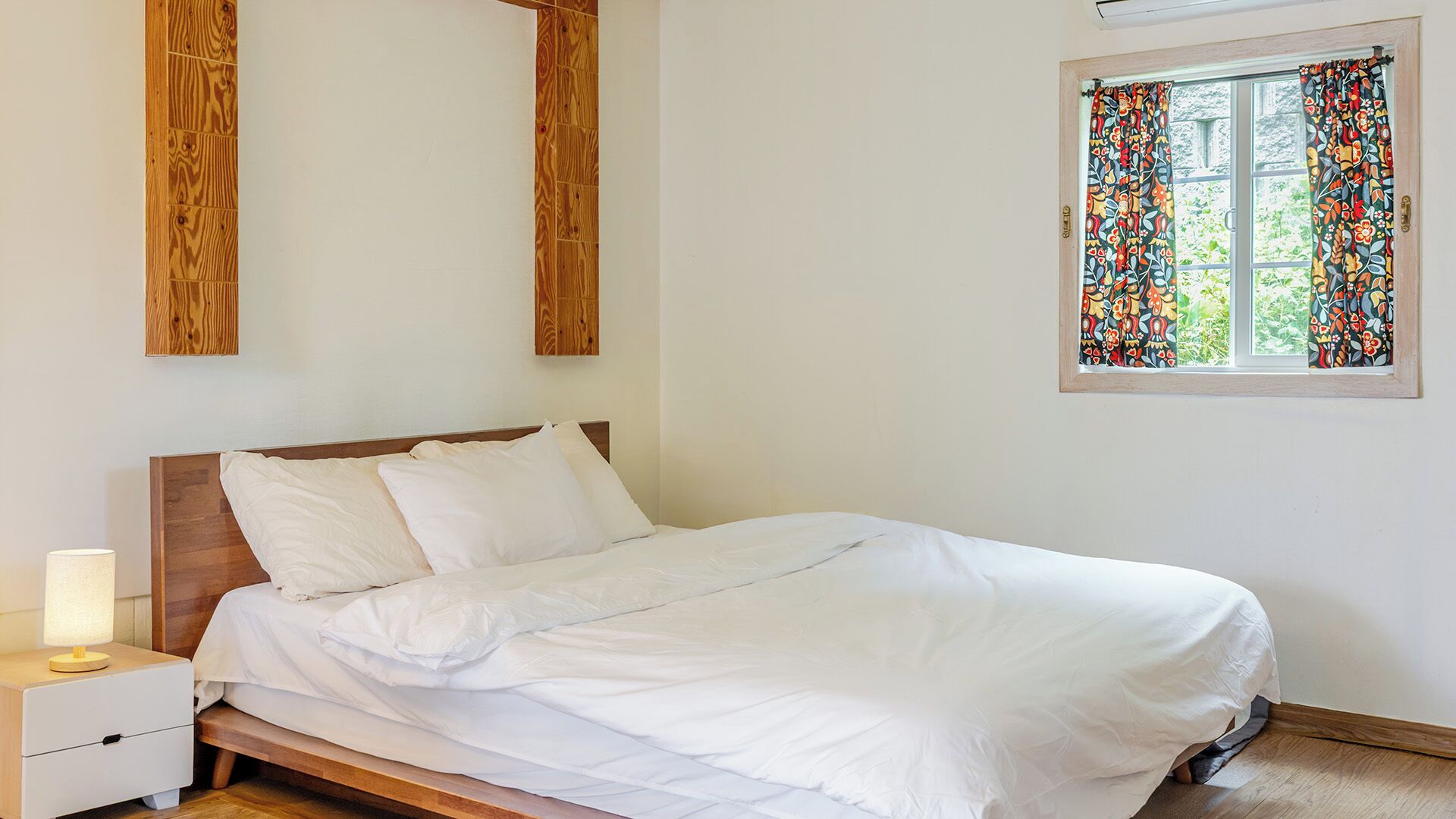 A bed with white bedding, a wooden headboard, a nightstand with a lamp, and a window with patterned curtains.