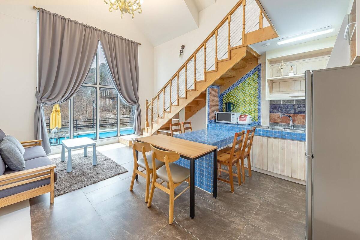 A modern living room with a sofa, dining table, and chairs. A staircase leads to a kitchen area with a tiled backsplash.