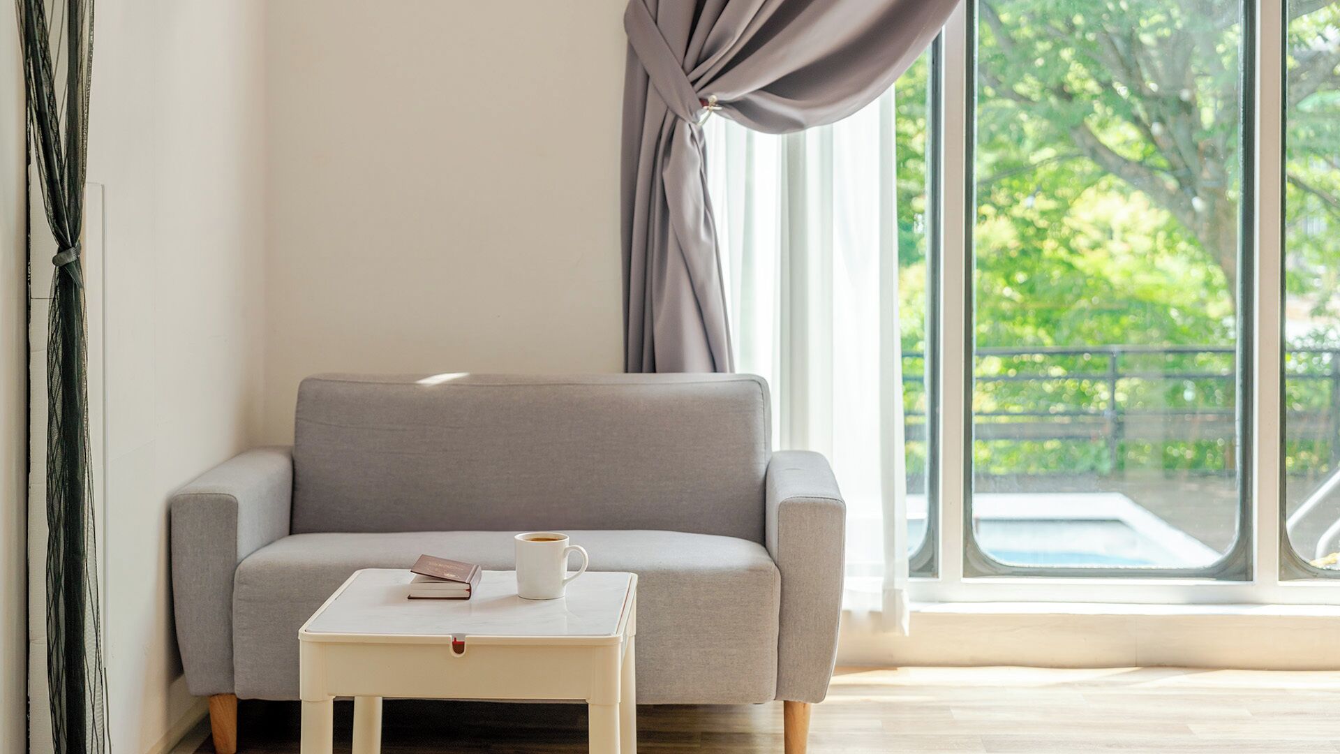 A modern living room with a grey sofa, a small white table with a book and a cup, and a large window with a view of greenery.