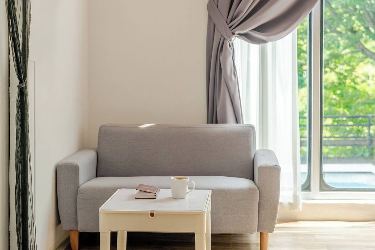 A modern living room with a grey sofa, a small white table with a book and a cup, and a large window with a view of greenery.