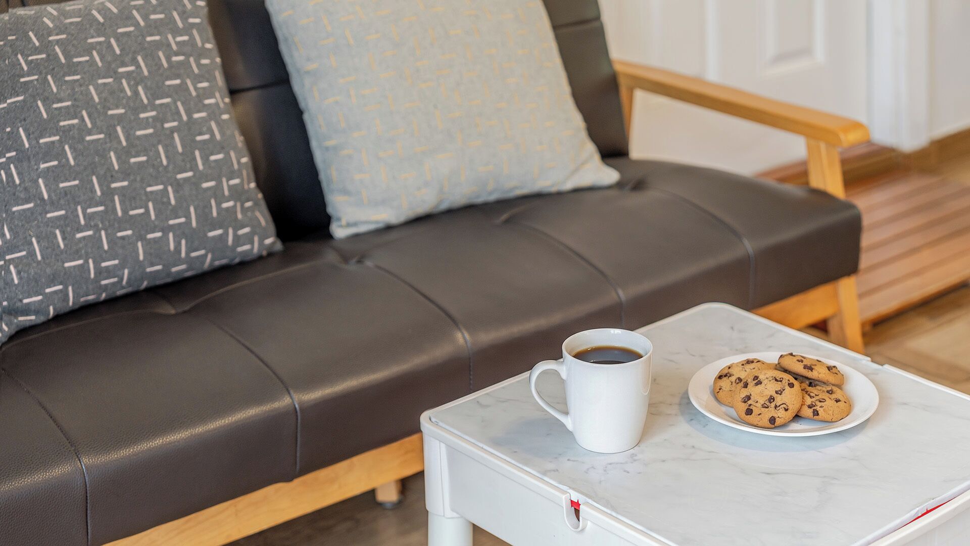 Ein braunes Ledersofa mit gemusterten Kissen, eine weiße Tasse mit Kaffee und ein Teller Kekse auf einem weißen Tisch.